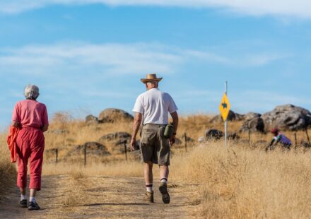 An older man and woman walking side by side on a dry dirt hiking trail under a blue sky