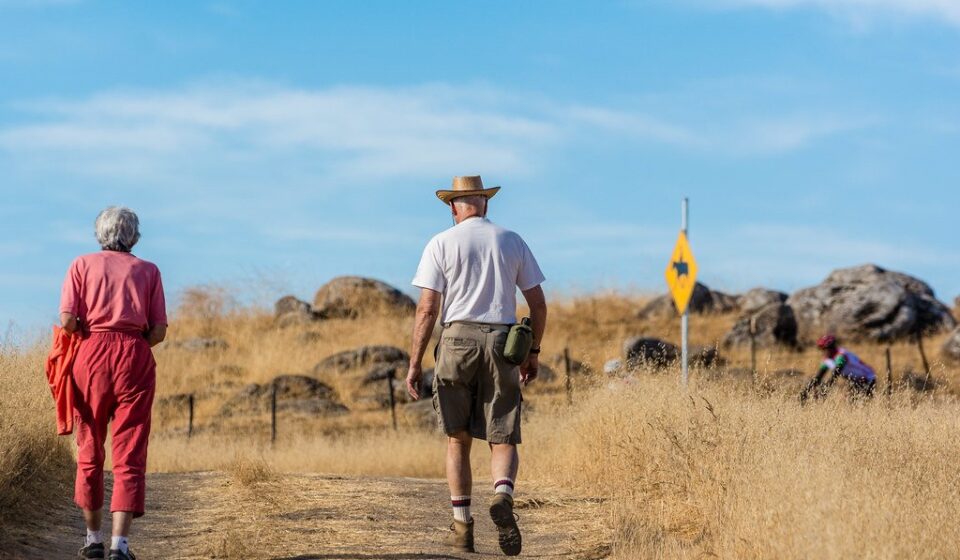 An older man and woman walking side by side on a dry dirt hiking trail under a blue sky
