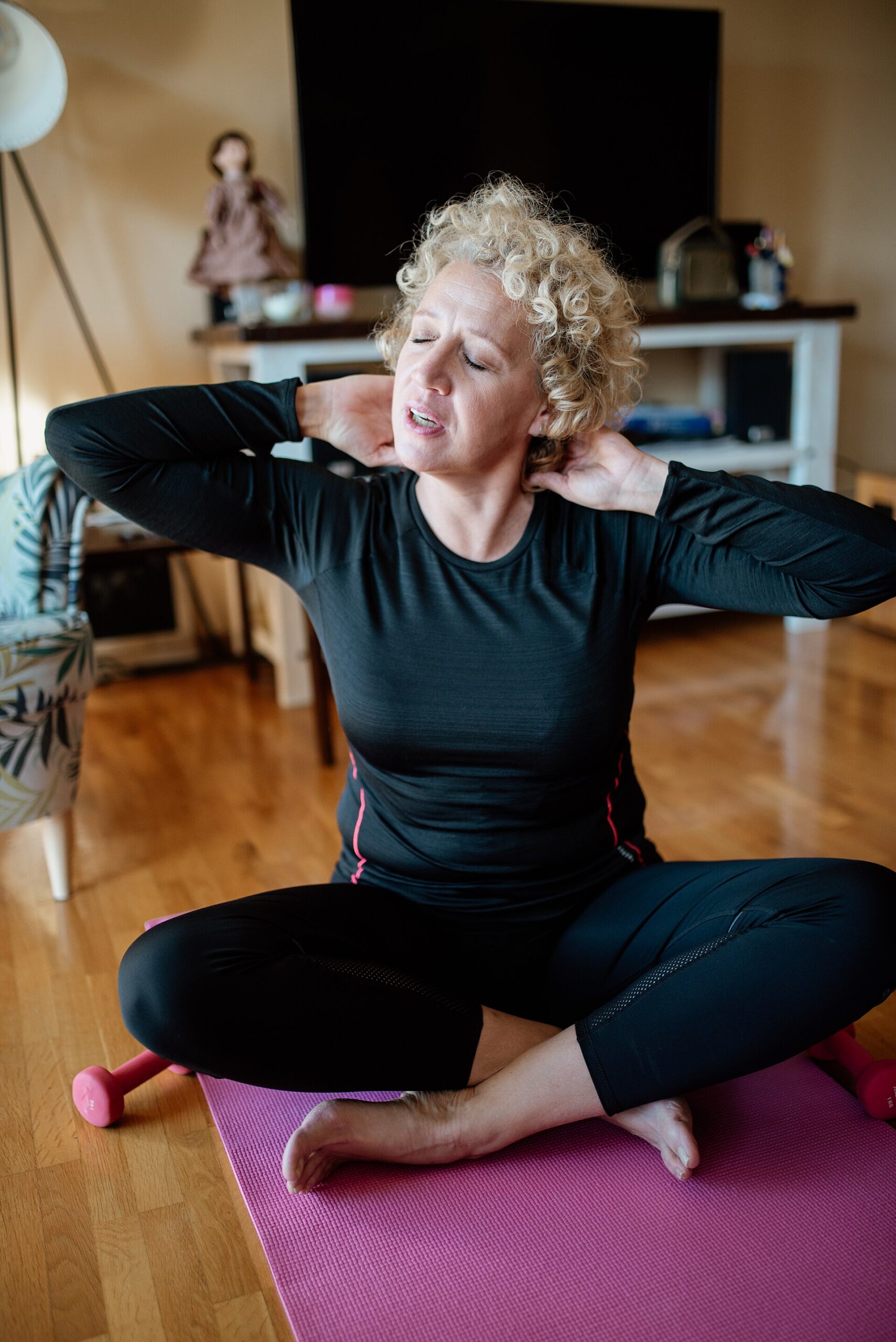 Woman with curly gray hair sitting cross-legged on a yoga mat at home, stretching her neck and shoulders