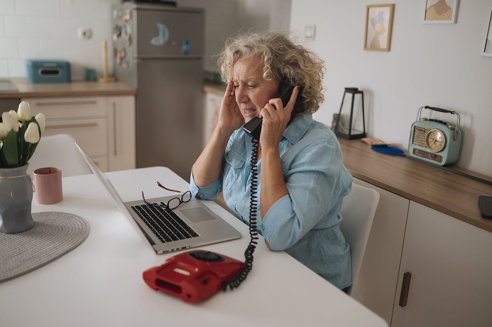 Older woman on the phone at her kitchen table with a laptop open