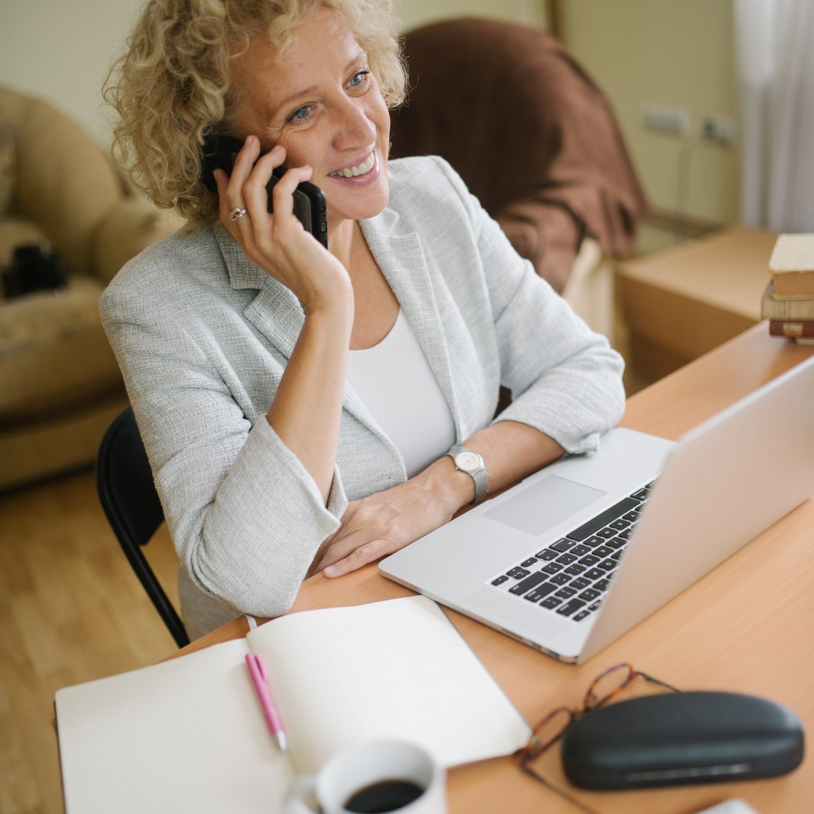 Senior woman talking on the phone at her desk while looking at notes and a laptop