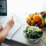 Person writing a meal plan at a kitchen table beside a bowl of fruit and a fresh salad