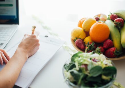 Person writing a meal plan at a kitchen table beside a bowl of fruit and a fresh salad