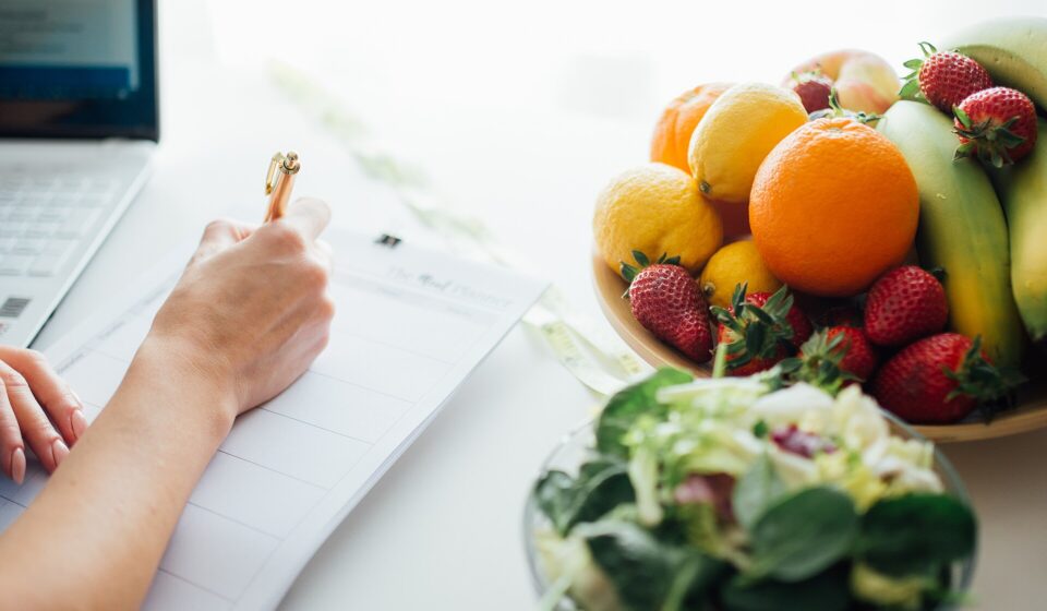 Person writing a meal plan at a kitchen table beside a bowl of fruit and a fresh salad
