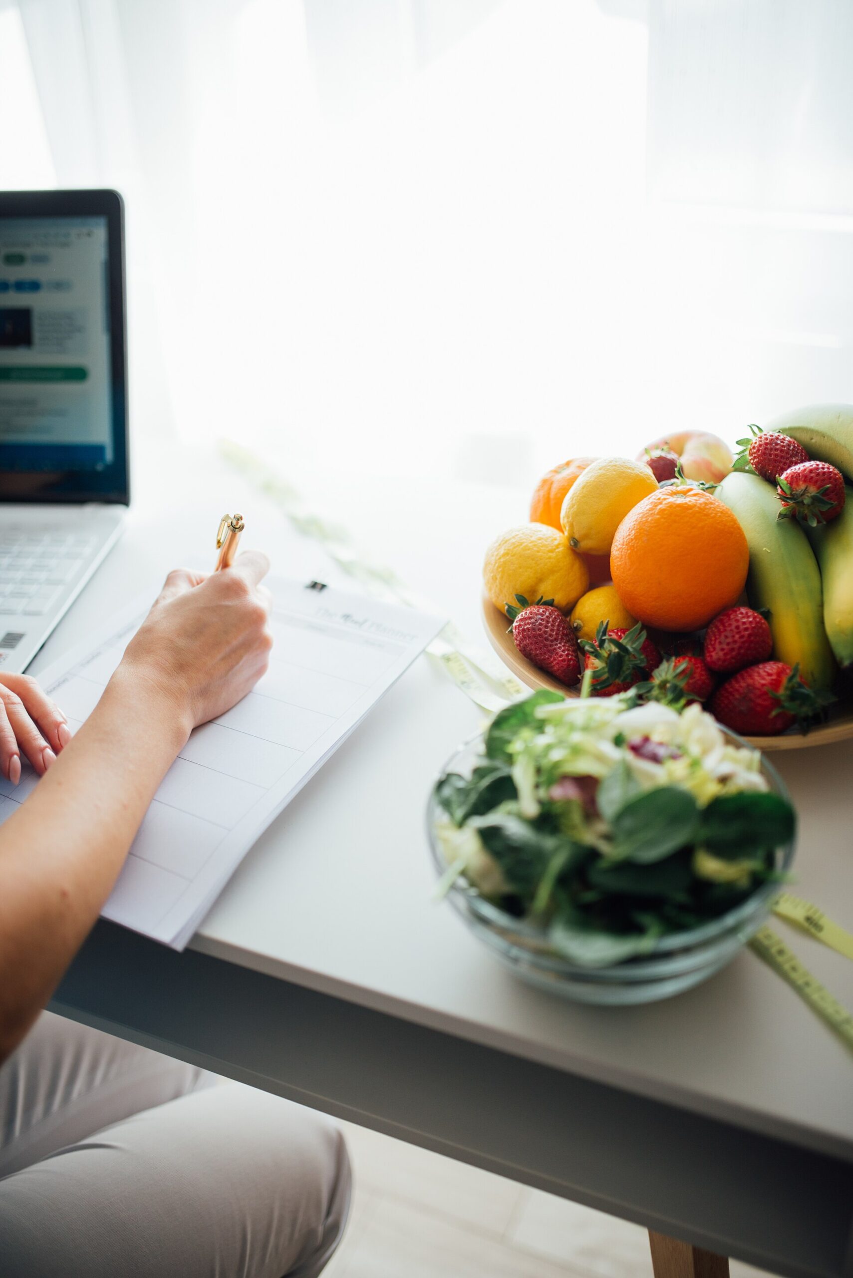 Person writing a meal plan at a kitchen table beside a bowl of fruit and a fresh salad