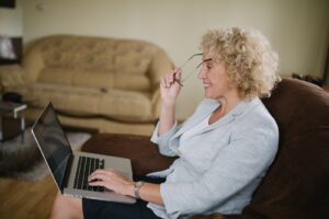 Senior woman with curly hair putting on her glasses while looking at a laptop