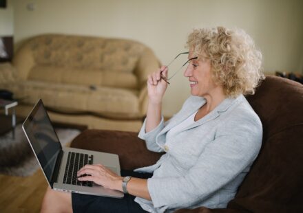 Senior woman with curly hair putting on her glasses while looking at a laptop