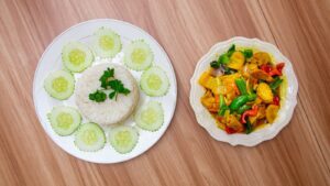 A plate of white rice with sliced cucumber next to a plate of mixed vegetables