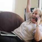 Senior woman with a laptop on her lap, talking on the phone and raising her arm in celebration