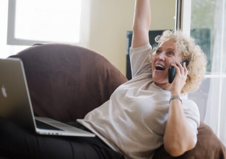 Senior woman with a laptop on her lap, talking on the phone and raising her arm in celebration