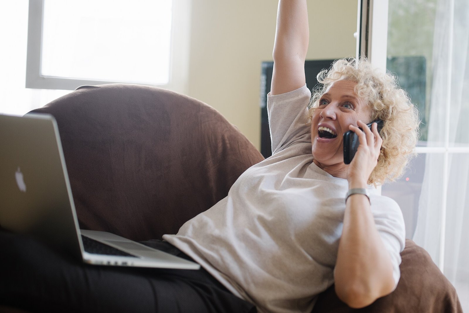 Senior woman with a laptop on her lap, talking on the phone and raising her arm in celebration