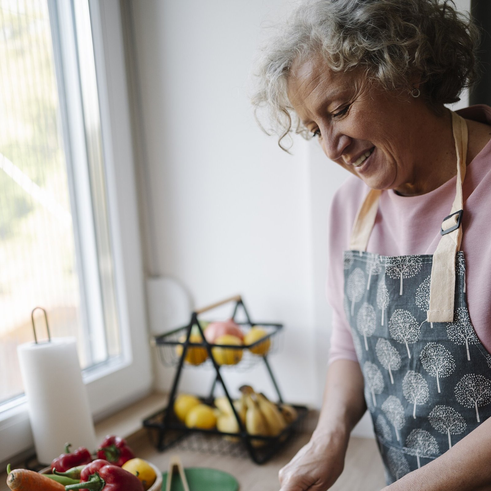 Senior woman smiling while slicing bread at a kitchen counter with fresh vegetables