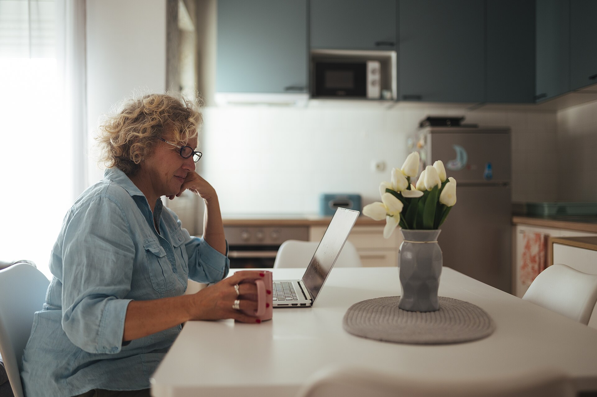 Older woman using a laptop in her kitchen with coffee and flowers