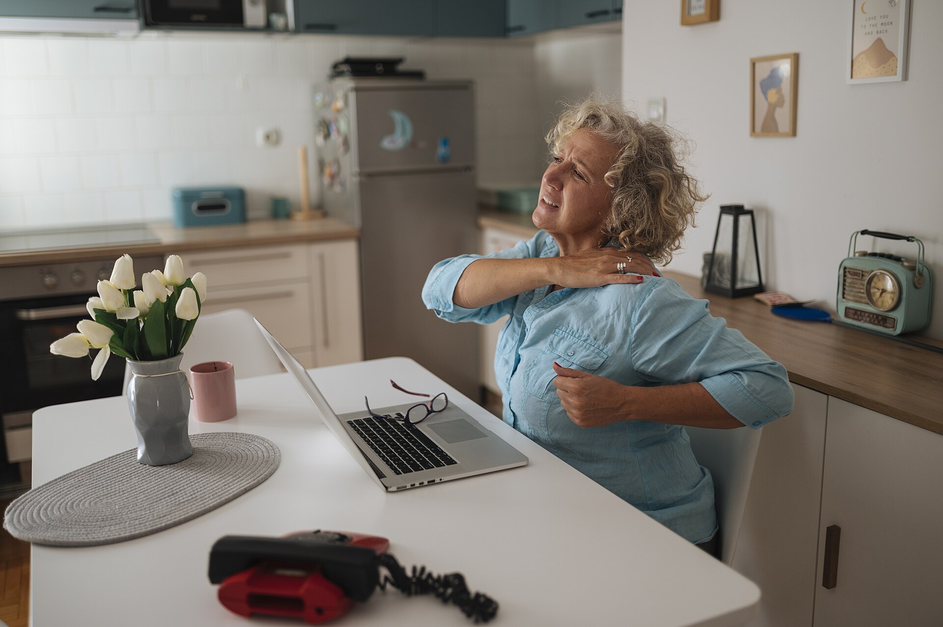 Older woman at a kitchen table rubbing a sore shoulder next to her laptop