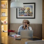 Older woman talking on the phone while sitting at a table with a laptop