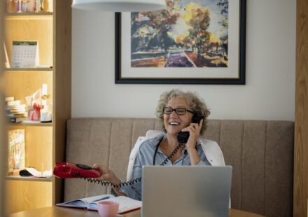Older woman talking on the phone while sitting at a table with a laptop