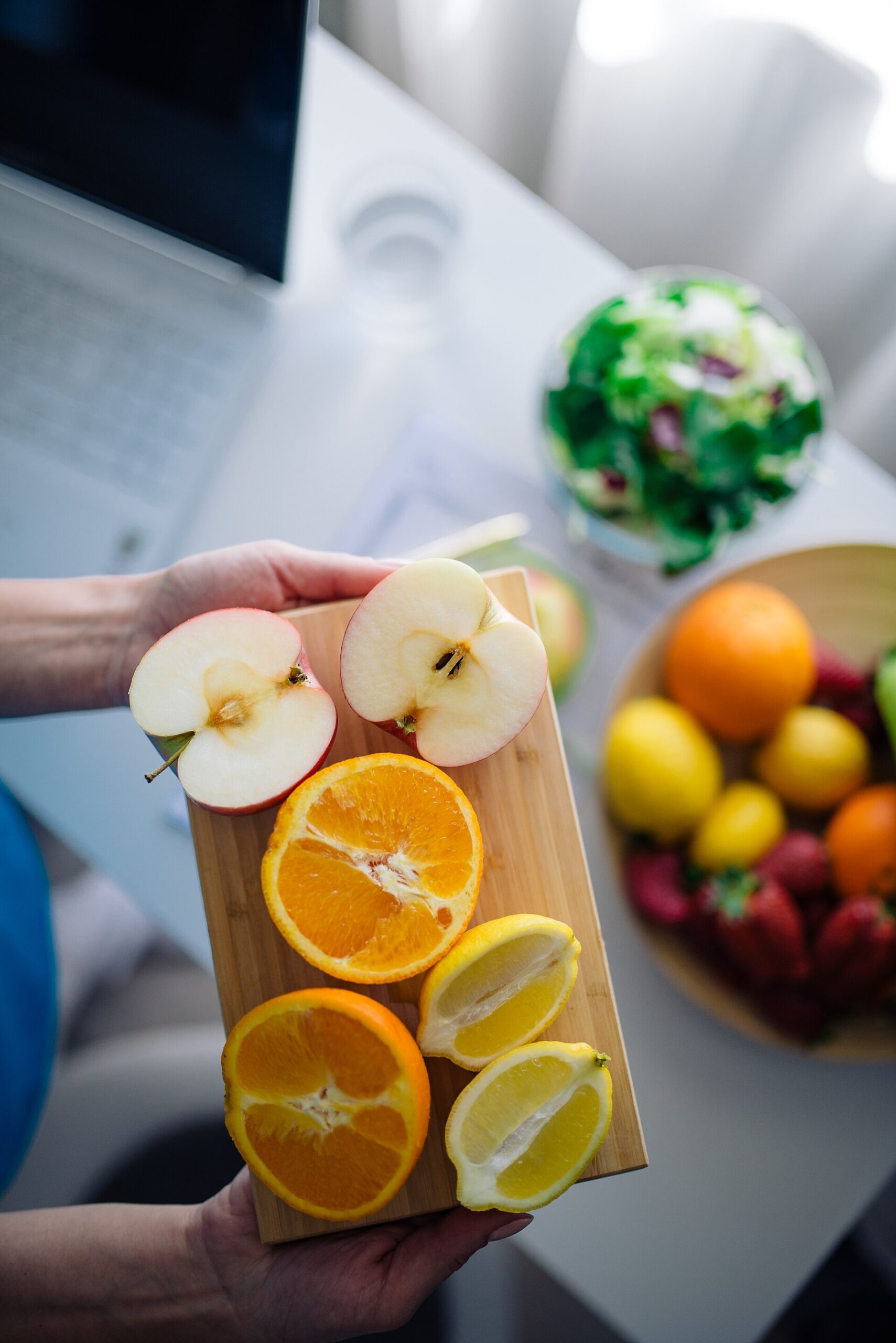 Hands holding a cutting board of sliced apples, oranges and lemons next to a bowl of salad