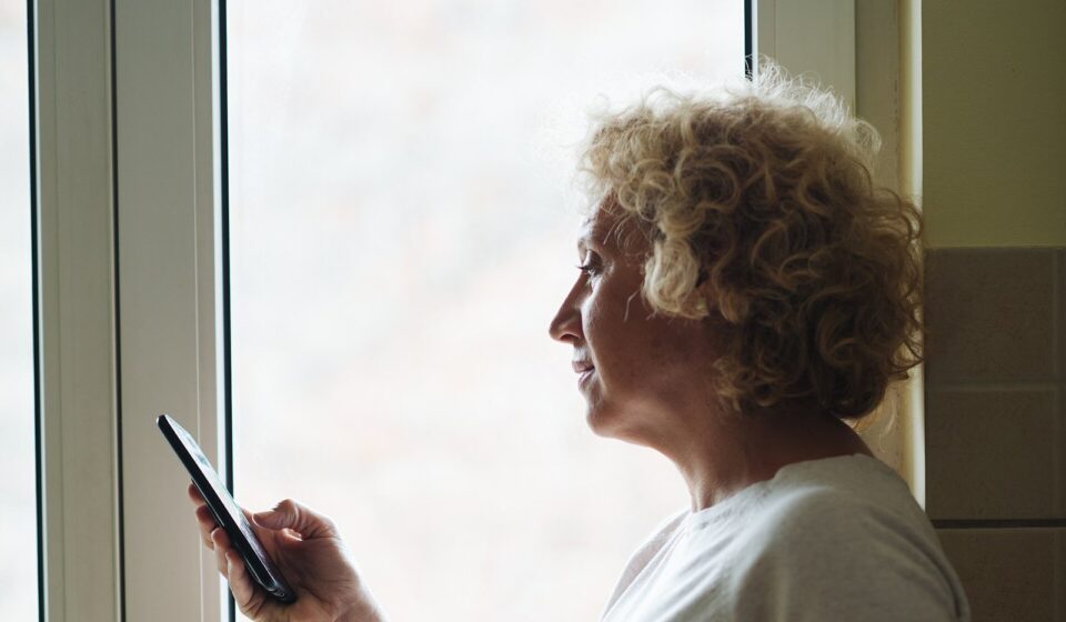 The $90 Medicare Part B Rebate: Who Gets It, When It Arrives, and What to Watch For Older woman standing by a window checking her phone